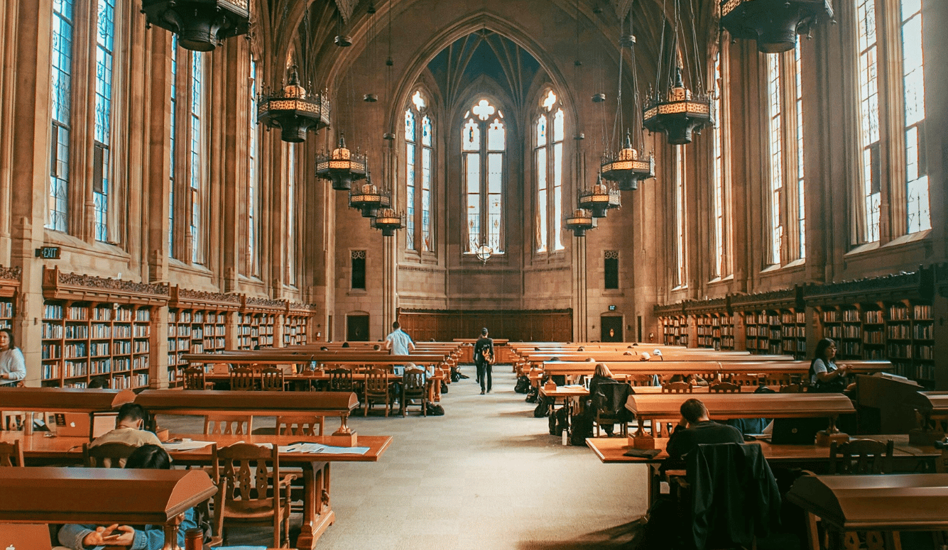 Students studying at long wooden tables in a Gothic university library with vaulted ceiling and stained glass windows. 