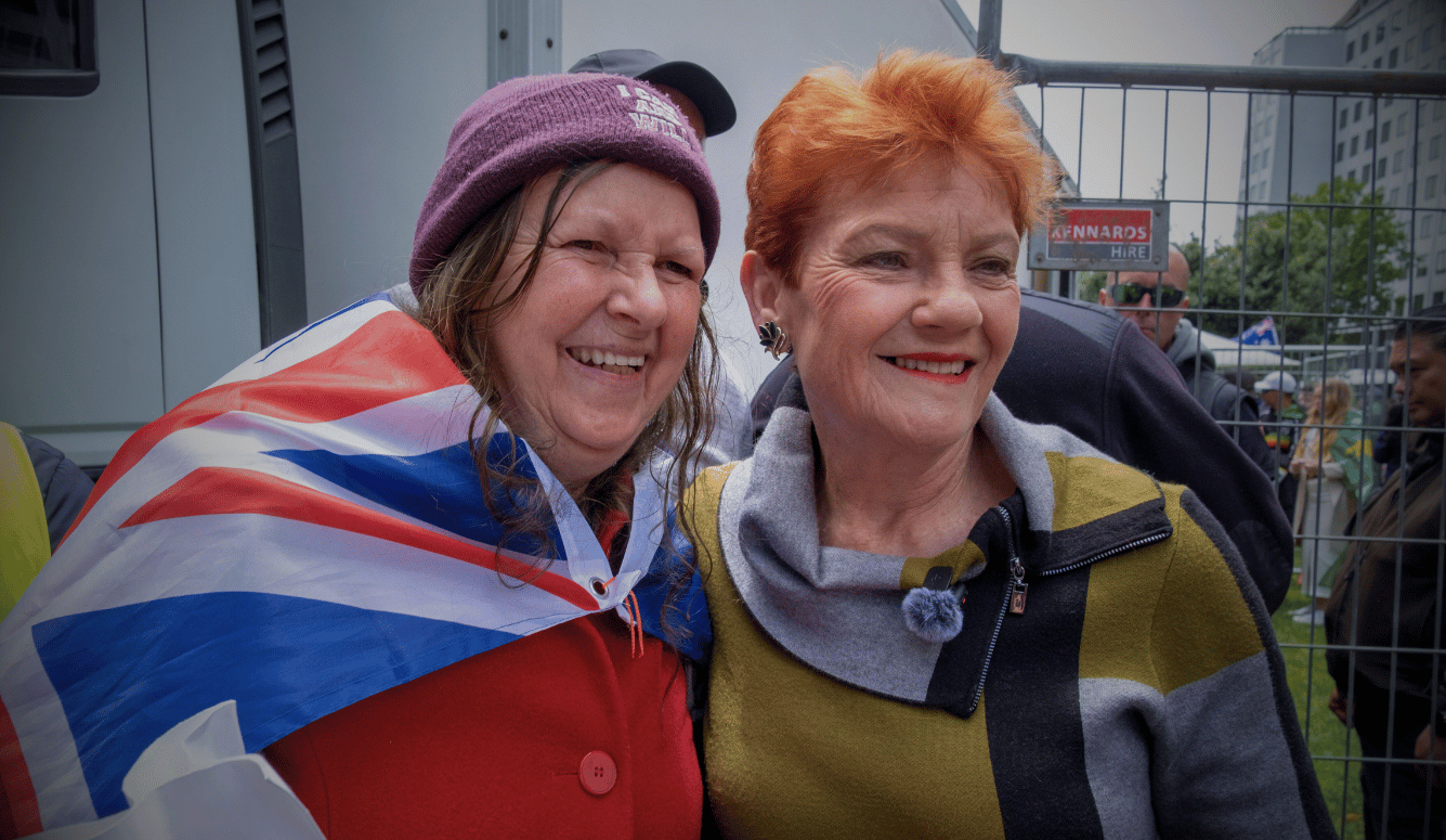 Pauline Hanson smiling with supporter draped in Australian flag behind security fencing at outdoor event. 