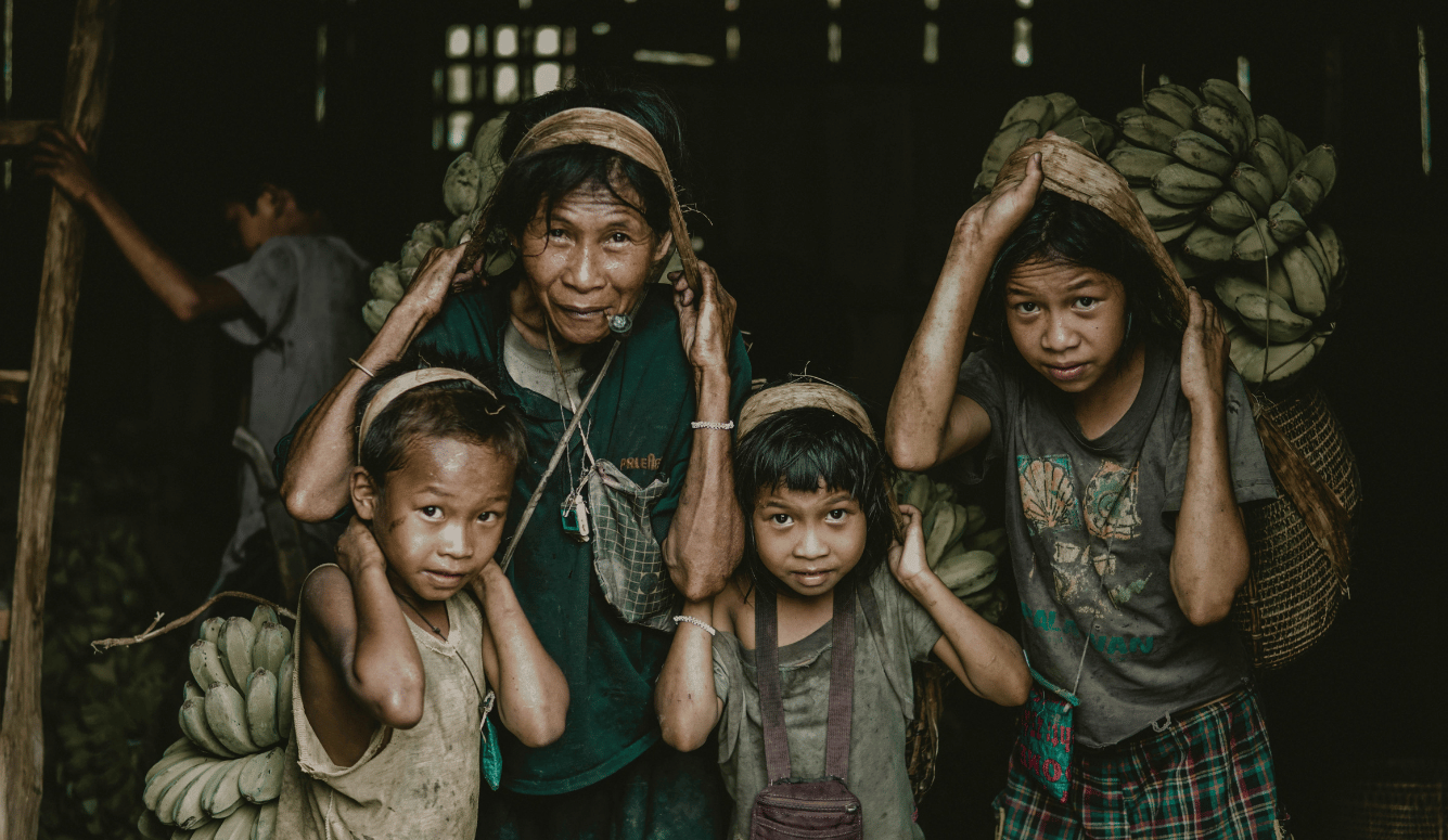 Four Filipino people, including an older woman and three children, carry banana baskets with head straps inside a rural shed.