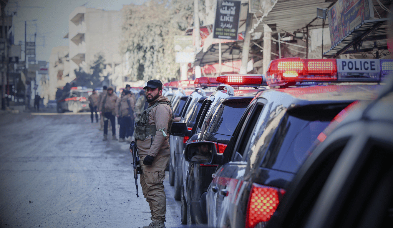 Syrian army officer holding a rifle beside parked police vehicles with flashing lights on a city street in Sheikh Maqsoud, January 2026.