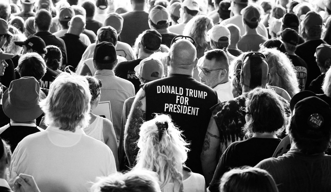 A crowd of people with their backs to us. One man sports a T-shirt with the slogan “Donald Trump for President.”