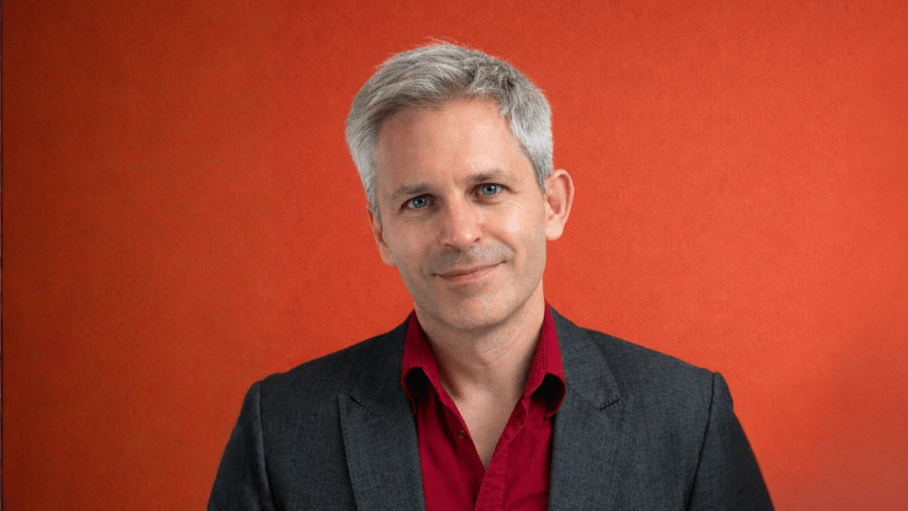Studio portrait of Shany Mor, a middle-aged man with short grey hair in red shirt and dark blazer against grey background.