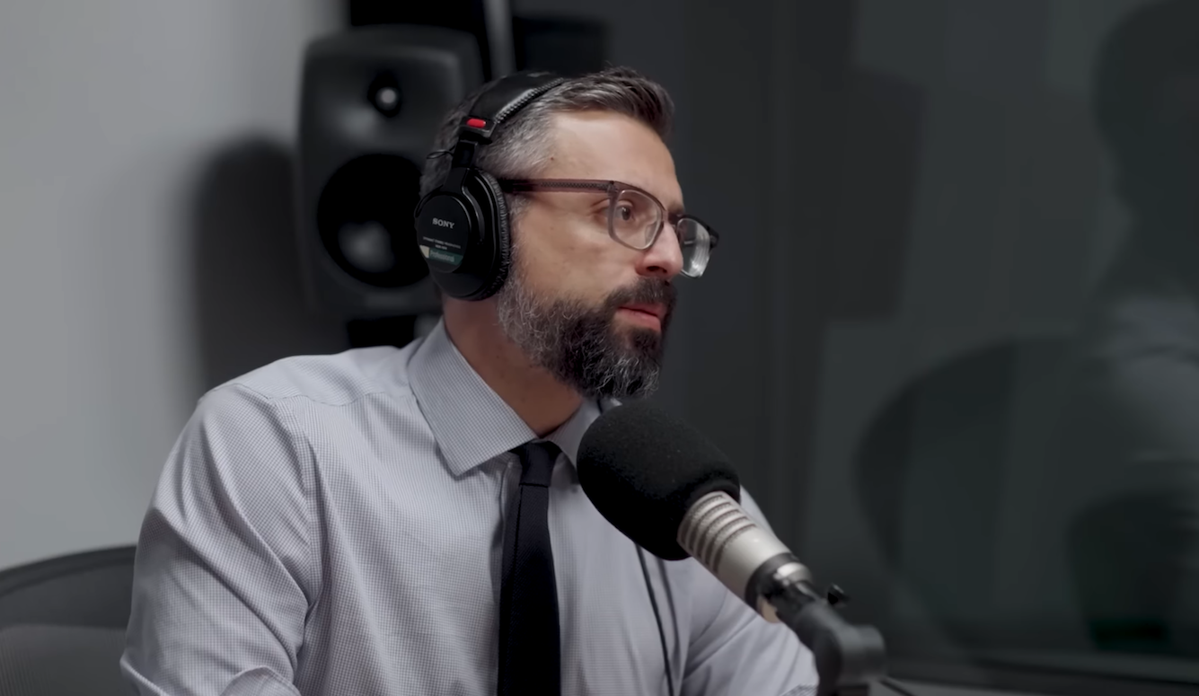 A man with glasses and a beard wearing headphones and a white collared shirt speaks into a professional microphone in a recording studio with monitor speakers in the background.