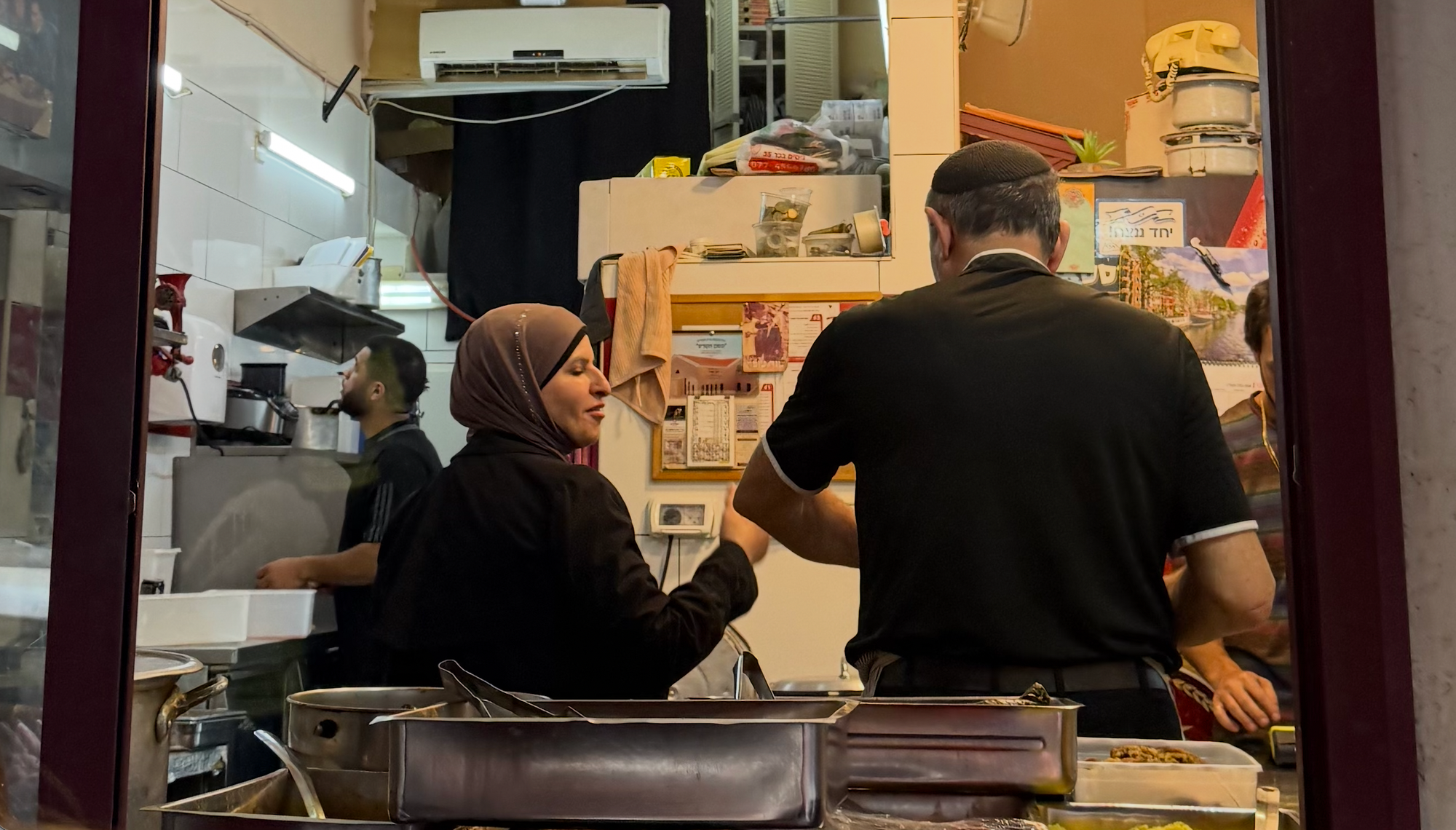 A Muslim woman in a hijab works in a falafel shop with a Jewish man wearing a kippah.