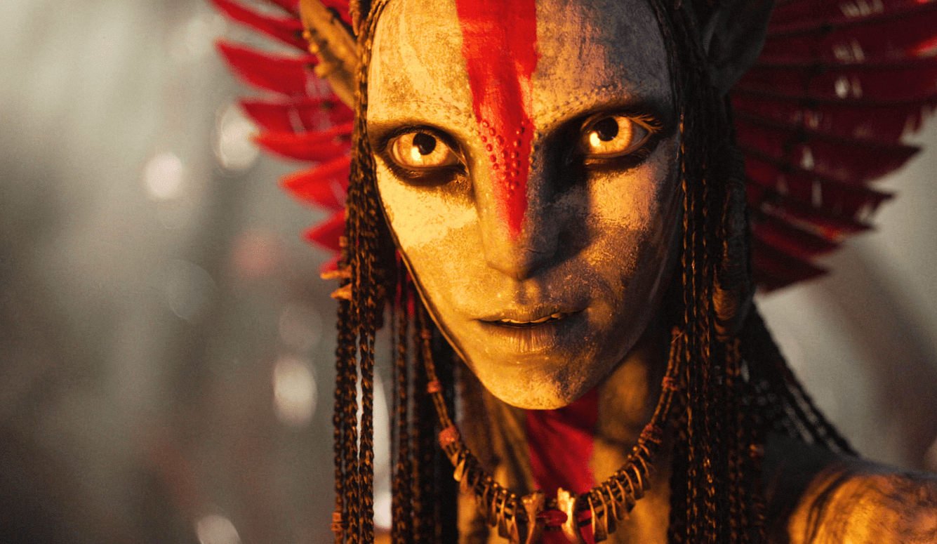 CGI humanoid figure with golden skin, red face paint, feathered headdress, glowing eyes, and beaded braids. Fantasy warrior aesthetic.