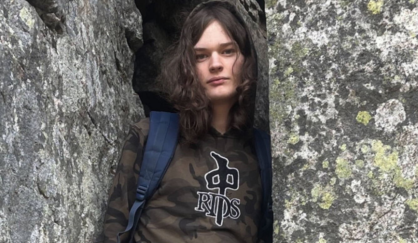 Young person with shoulder-length wavy hair wearing a camouflage hoodie with "RIPS" logo and blue backpack, standing against textured granite rock face with lichen patches.