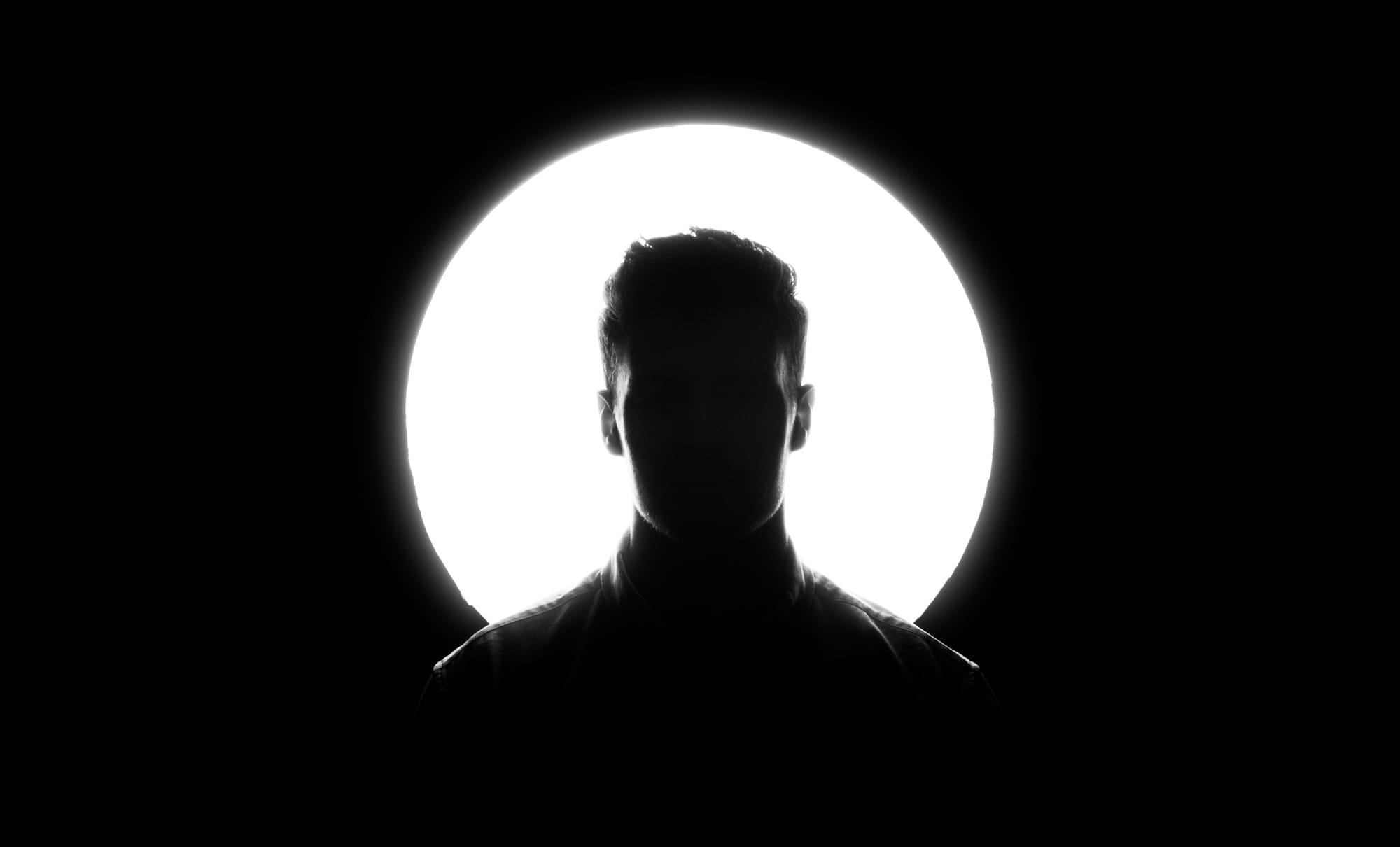 Silhouette of short-haired man in jacket against bright circular light on black background. 