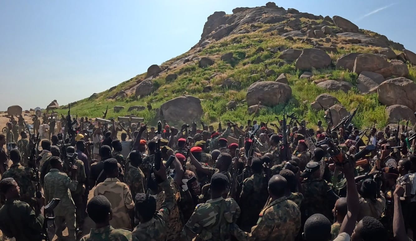 A large group of armed soldiers in camouflage and red berets raise their weapons in the air in front of a rocky, grass-covered hill under a clear blue sky.