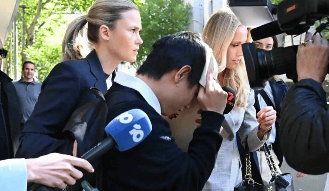 A young Asian man covers his face with a cloth while being escorted through a media pack outside a court building, surrounded by microphones and cameras.