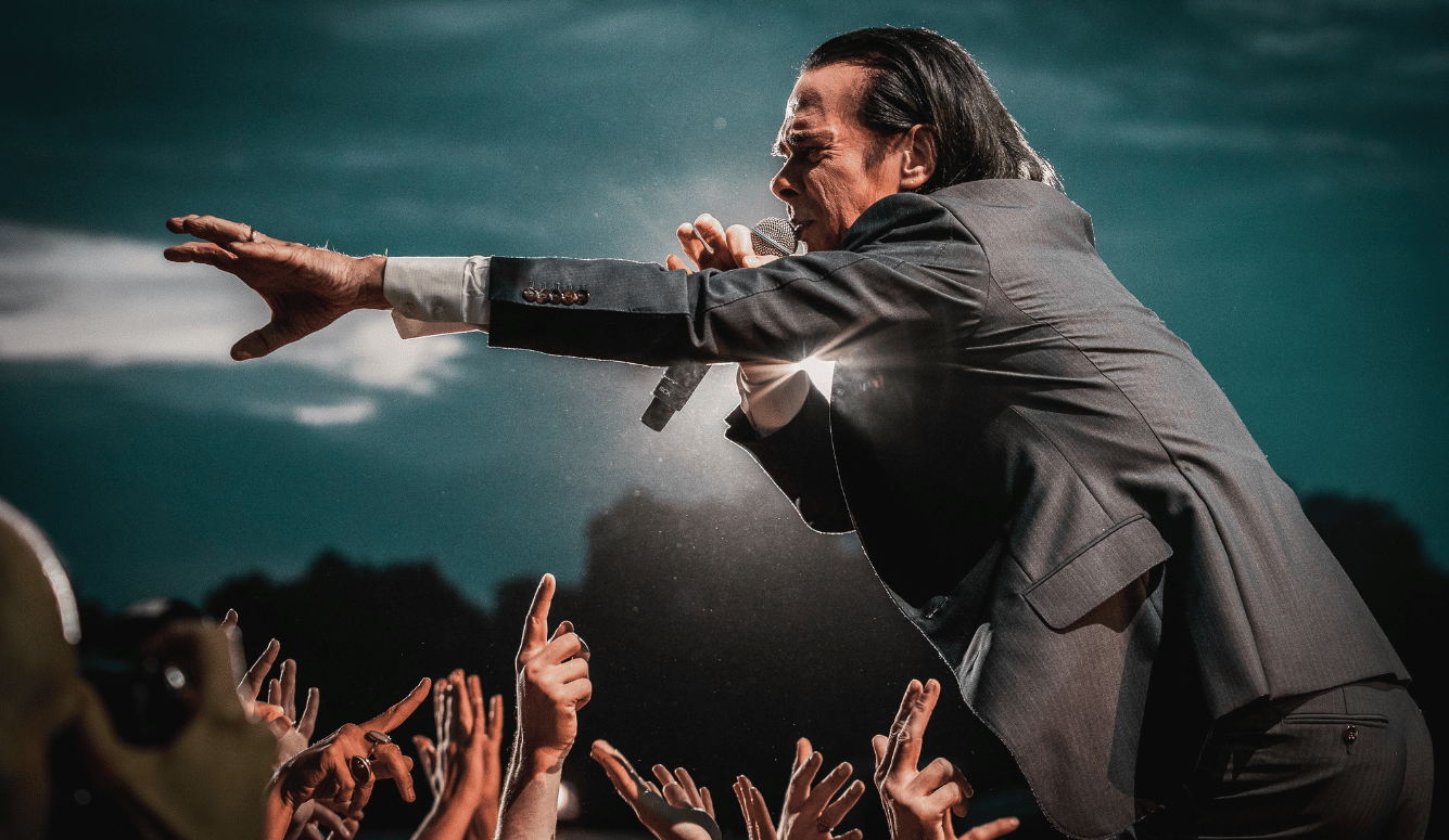 Nick Cave performing live, holding microphone and reaching towards raised audience hands at All Points East Festival, London, 2022.