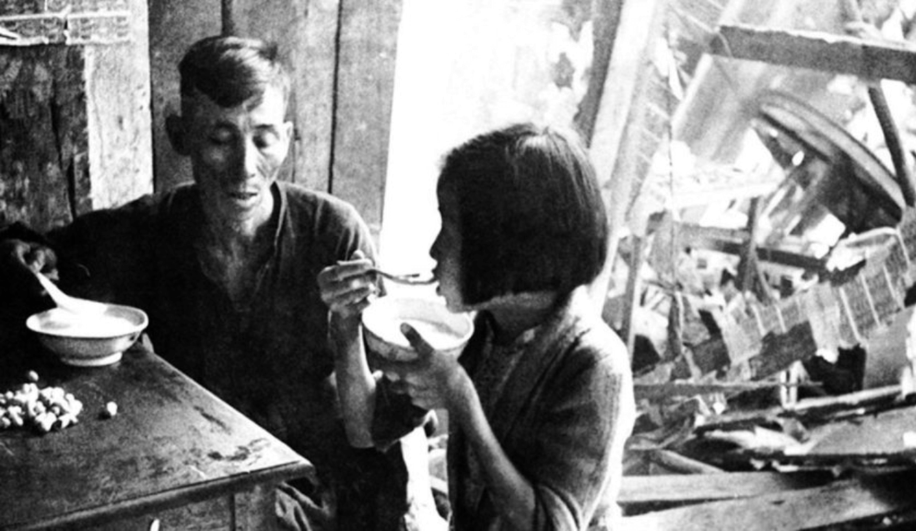 A black-and-white photo of a man and a young girl eating meager rations from small bowls while sitting amidst the rubble of a destroyed building.