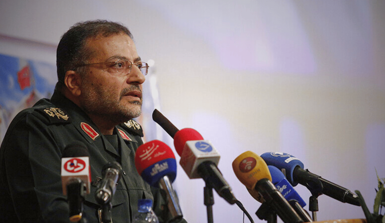 Arab man in military uniform and glasses speaks into many microphones.