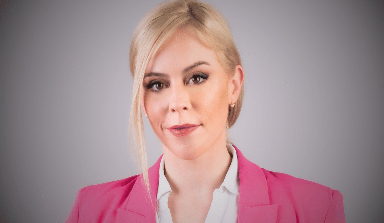 Emily Schrader has blonde hair, wearing a pink blazer and white blouse, with a composed and professional expression.