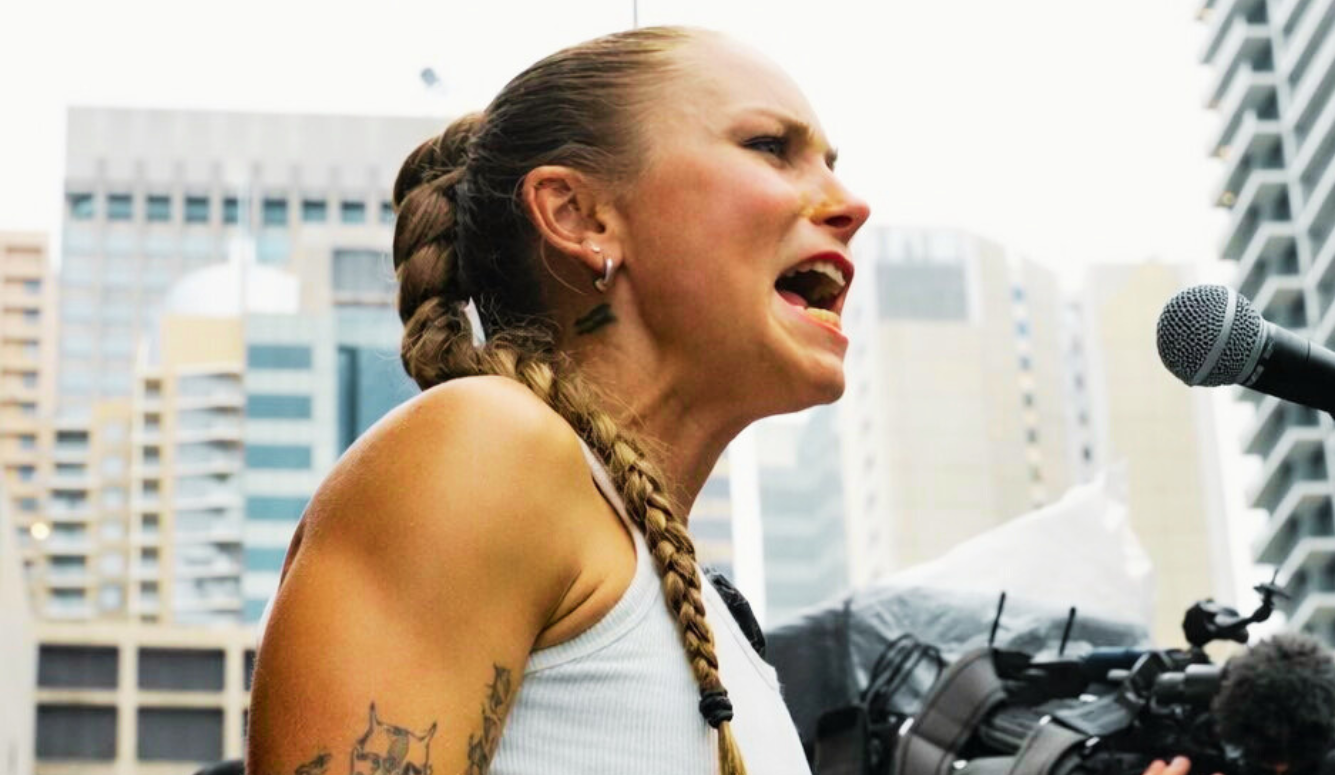 Woman with French braids speaks passionately into a microphone at an outdoor urban rally, with city buildings in the background.