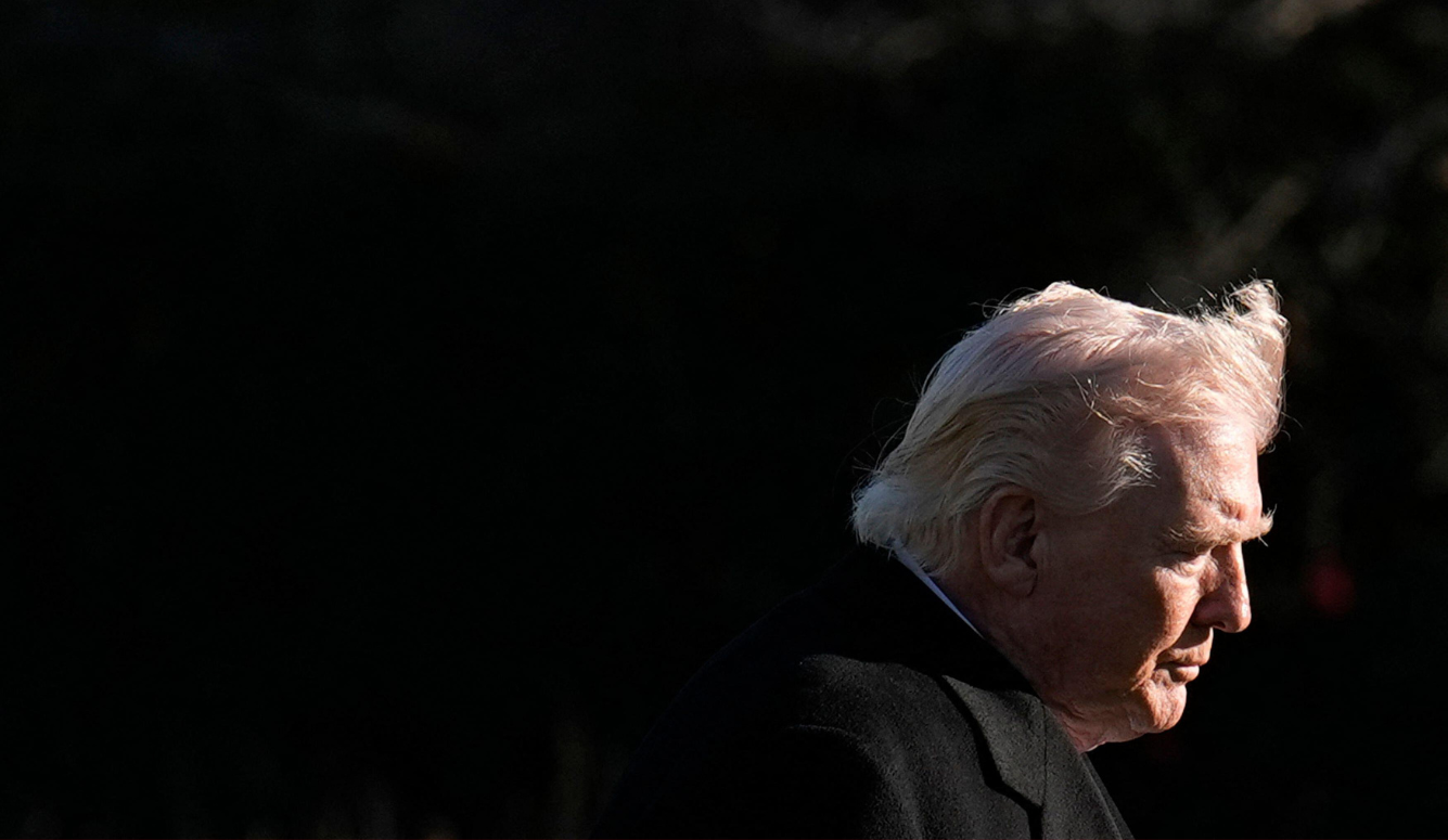 Donald Trump's head and shoulders are visible as he walks in a breeze. Dark jacket, dark background. 