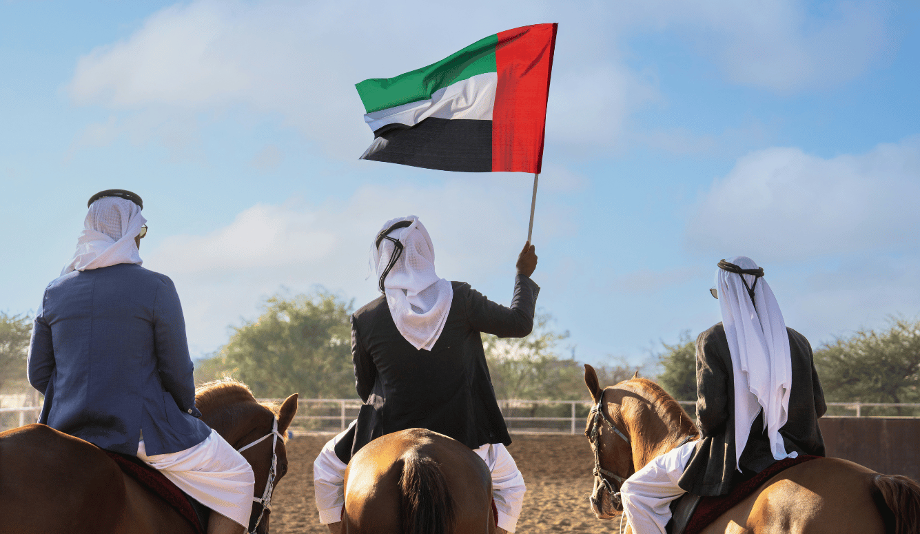 Three Arab men on horseback, viewed from behind, in a paddock. The middle one holds up a UAE flag. 