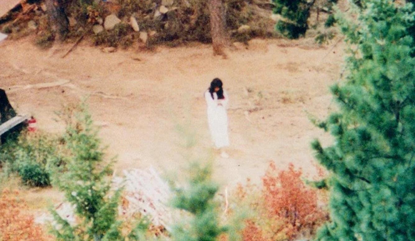 A photograph of a young woman in a forest clearing. She wears a white dress and sneakers and has long, dark hair. 