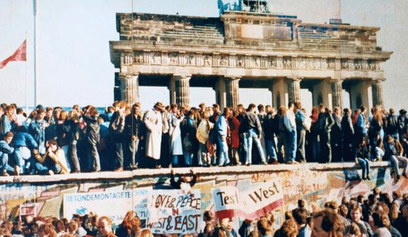 Crowds on top of and in front of the Berlin Wall, some carrying banners. 