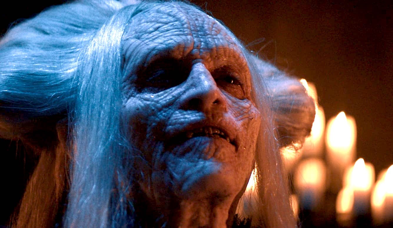 Pale blue-skinned creature with heavily wrinkled face, white hair, and exposed teeth, lit by candlelight; fantasy/horror film prosthetic makeup.