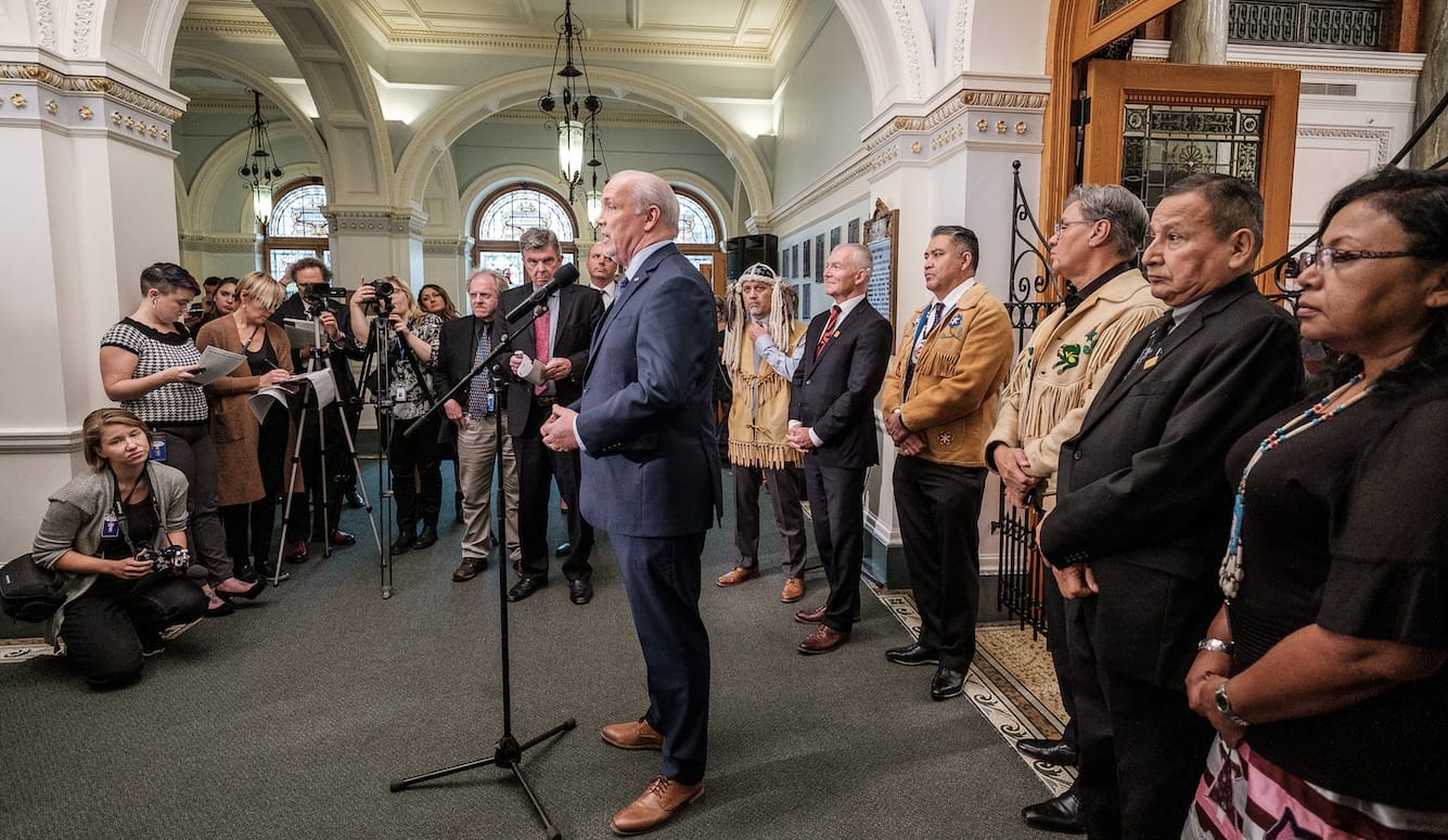 A man in a navy suit speaks at a microphone stand in an ornate government building rotunda, flanked by Indigenous leaders in traditional dress, while press photograph the event.