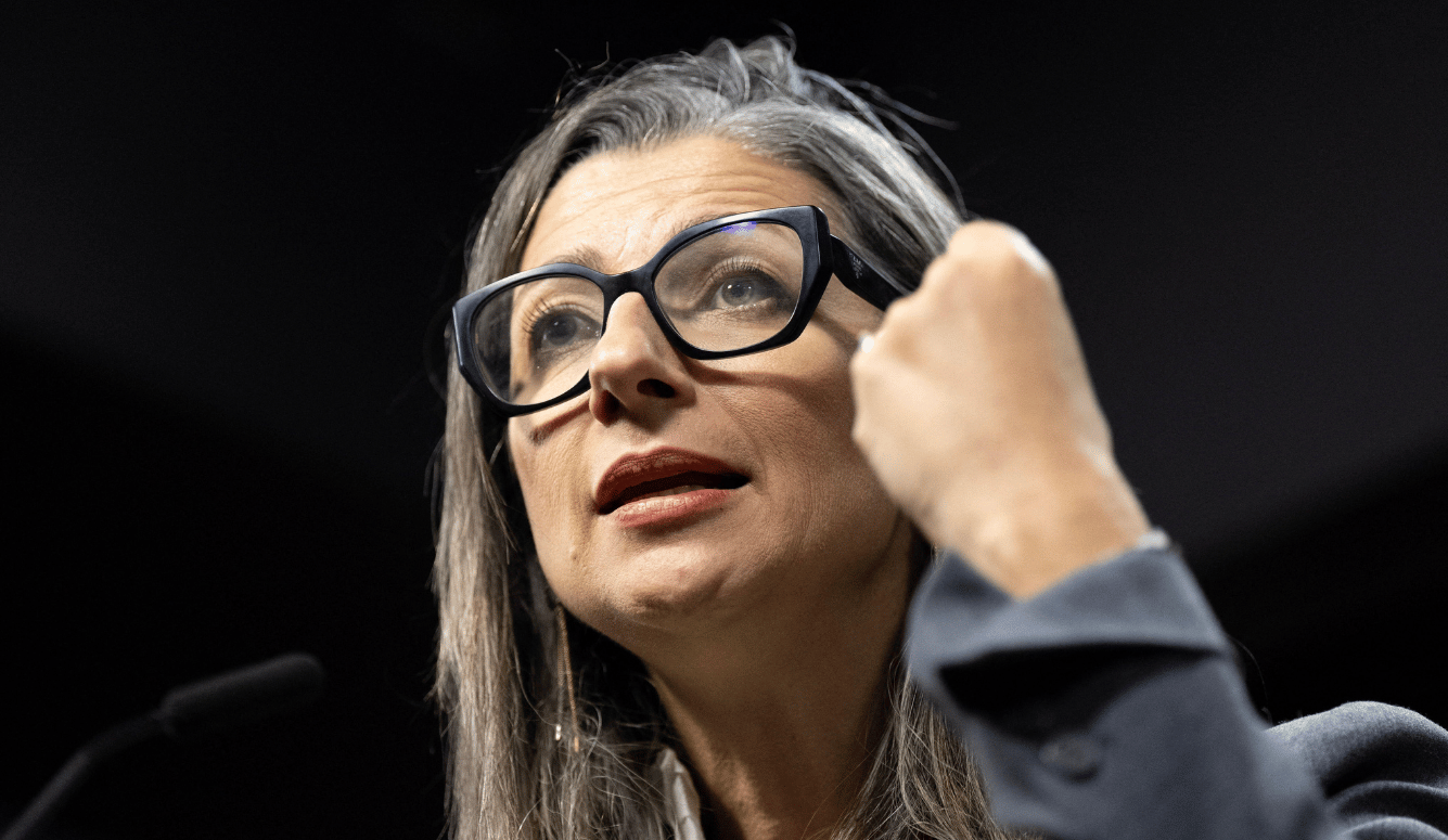 Woman with grey-streaked hair and black cat-eye glasses speaks passionately, raised fist gesturing, dark background, grey blazer.