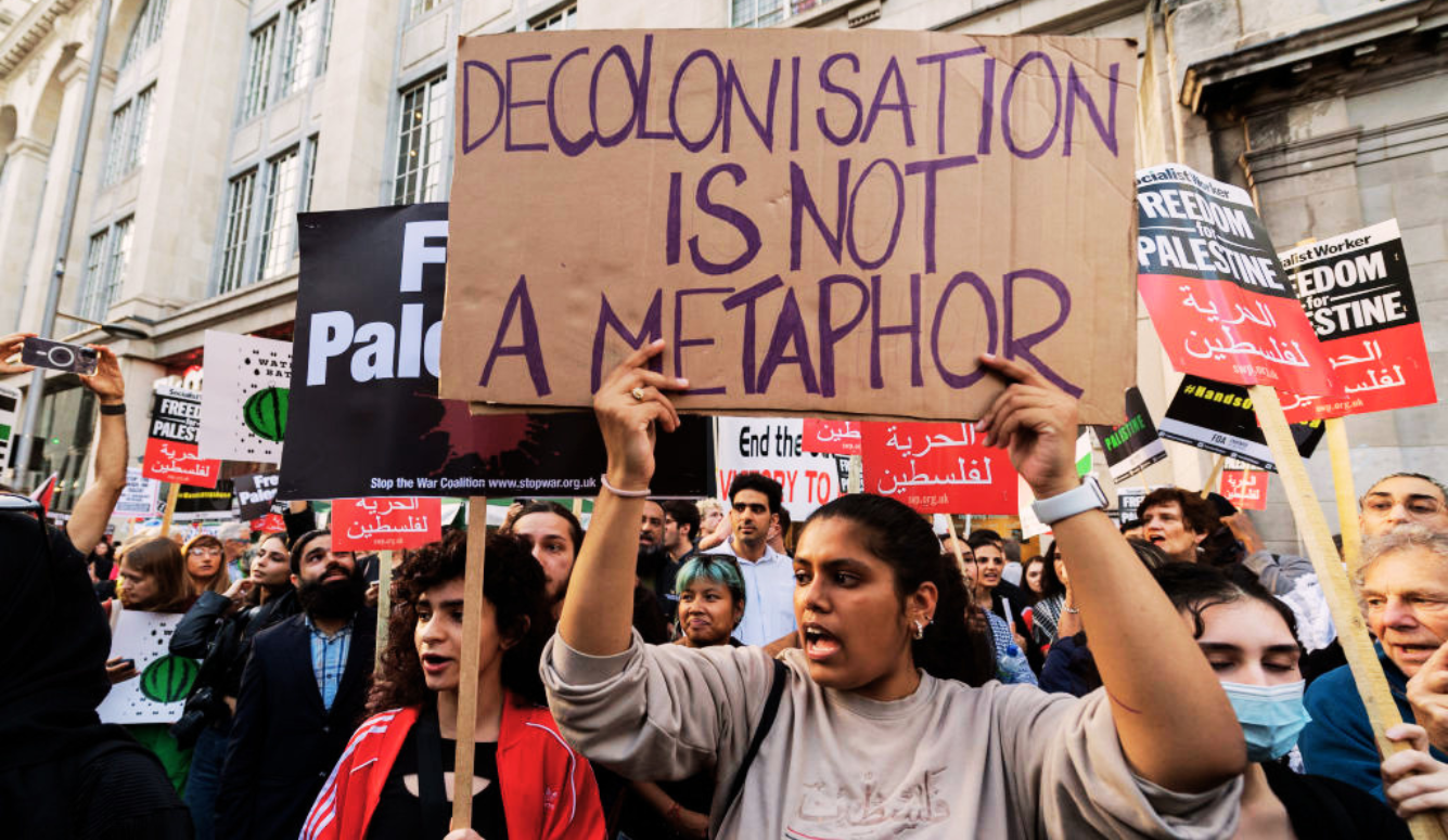 A woman at a protest holds a sign saying "Decolonisation is not a metaphor."