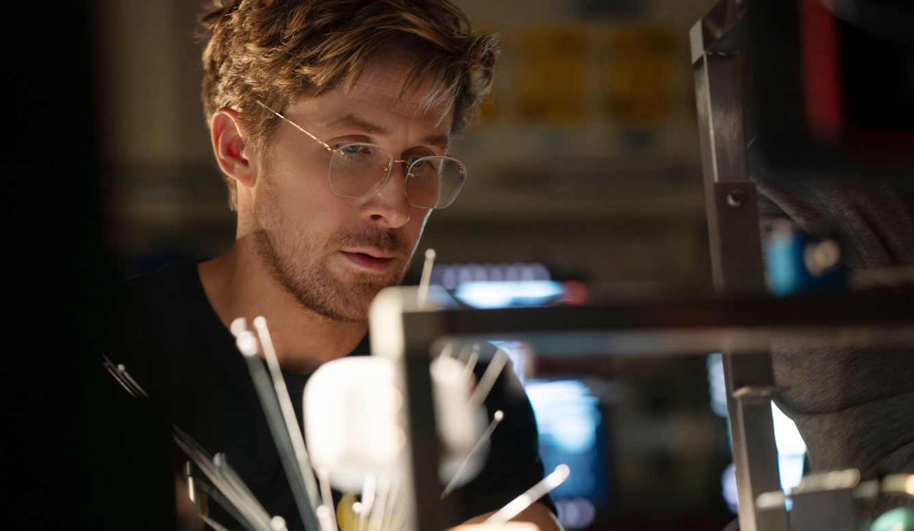 A man wearing wire-rimmed glasses examines a complex metal and wire apparatus in a dimly lit lab, his expression focused and intent.