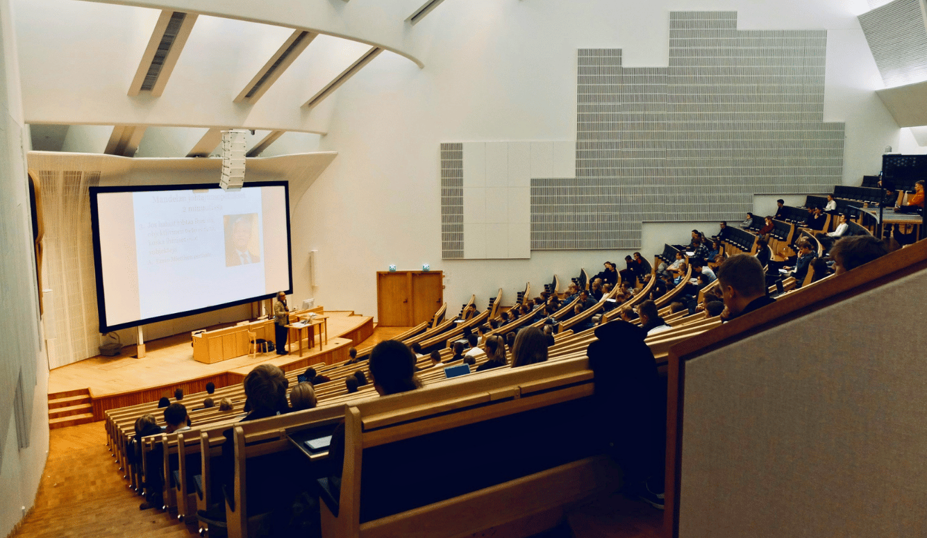 University lecture hall with tiered seating, students attending a presentation. A slide with text and a portrait is visible on the large screen at the front.