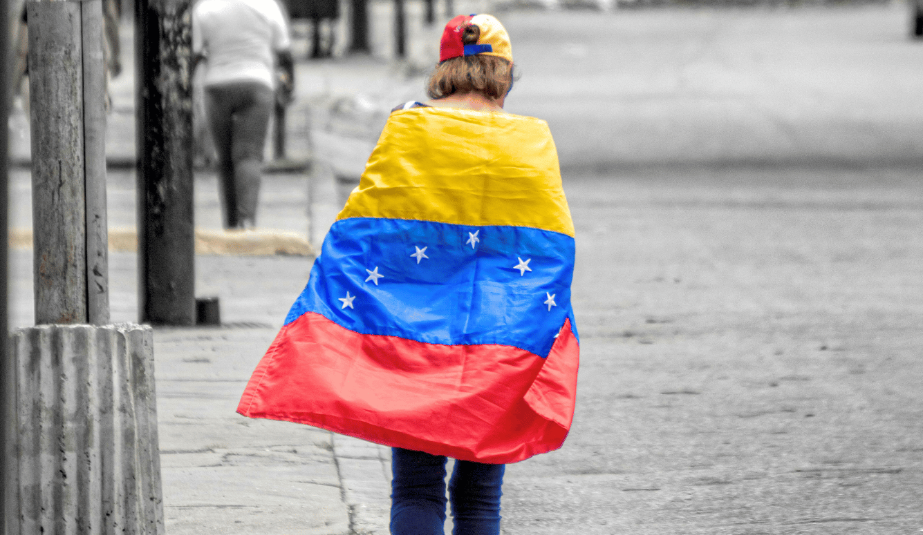 Person walking away draped in Venezuelan flag, wearing matching cap, on a grey city street. Selective colour highlights the flag's yellow, blue and red against monochrome background.
