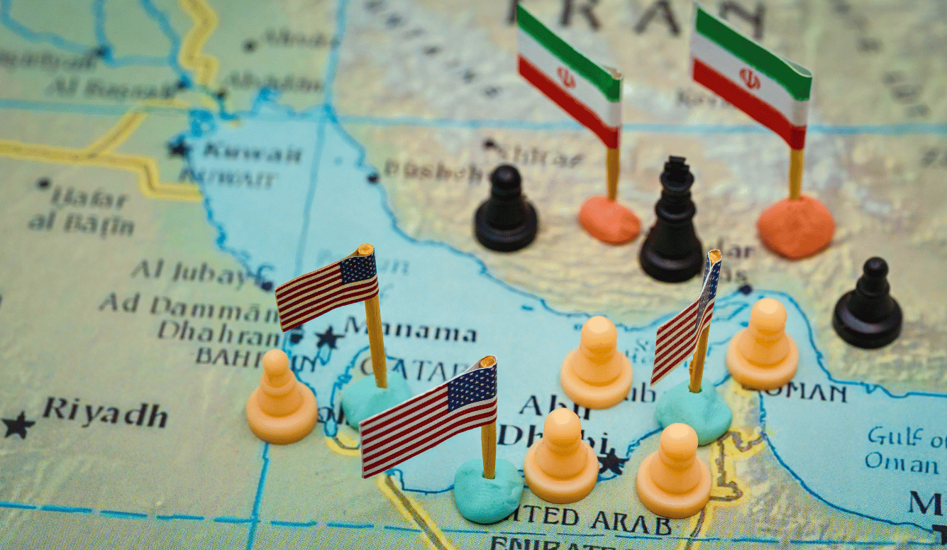 US and Iranian flags on miniature chess-piece stands placed on a map of the Persian Gulf region, symbolising geopolitical tension over the Strait of Hormuz.