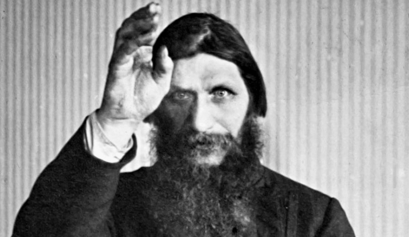 Black and white image of Grigori Yefimovich Rasputin. He is heavily bearded and is raising a hand, looking straight at the viewer.