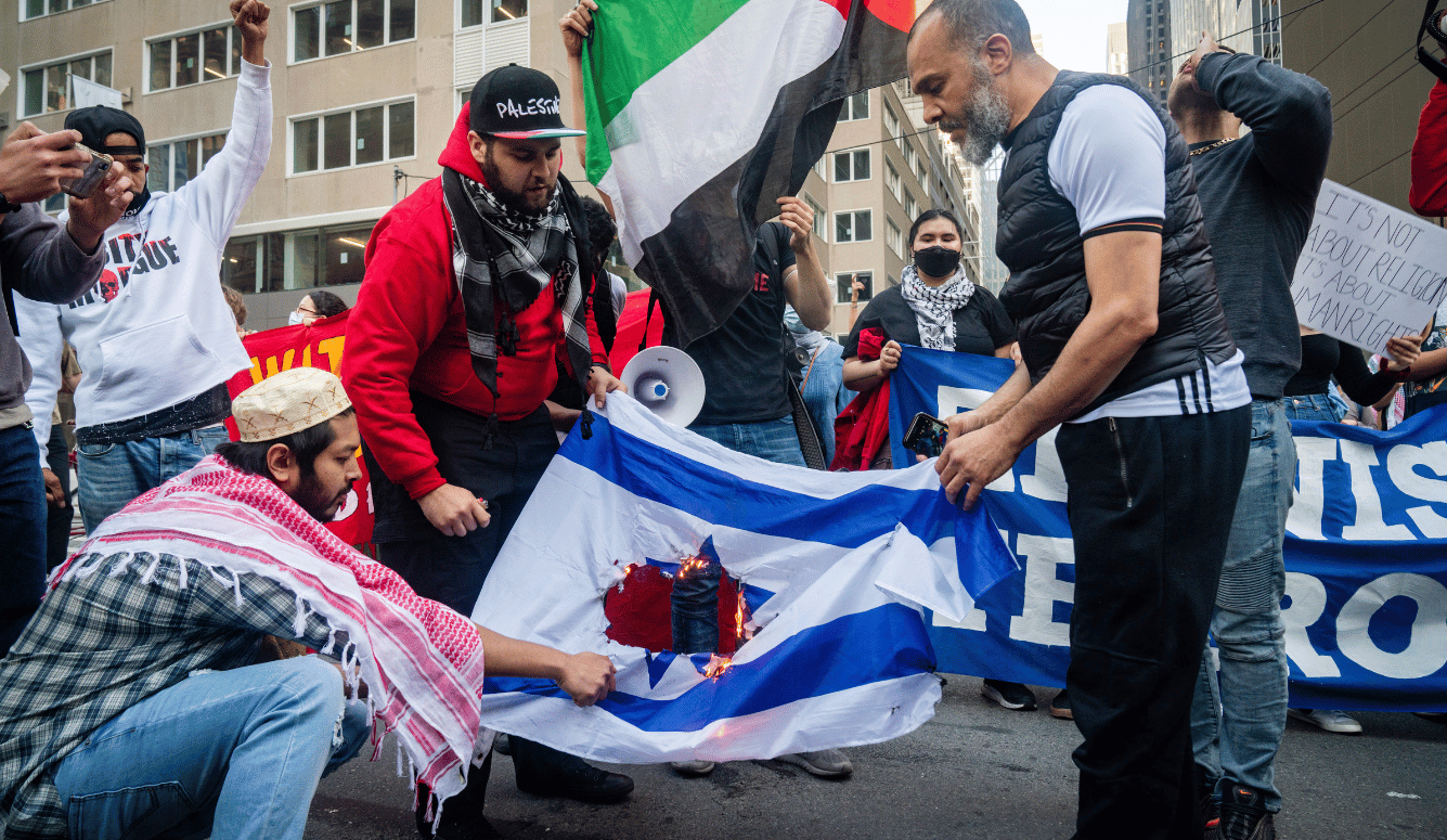 Protesters burning an Israeli flag while holding a large Palestinian flag during a street demonstration.