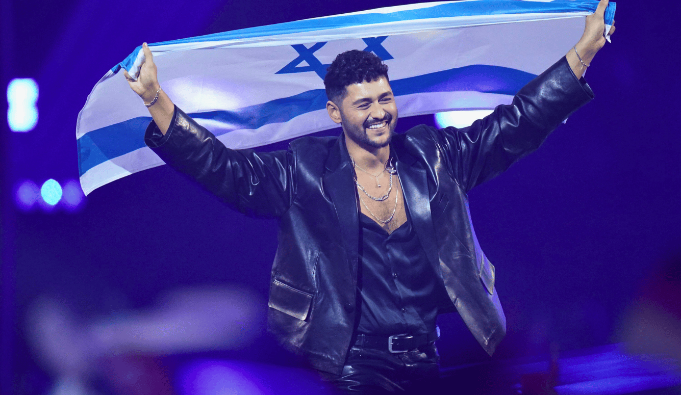 Young man of Middle Eastern appearance, Noam, in leather jacket, smiles as he holds the Israeli flag on stage.