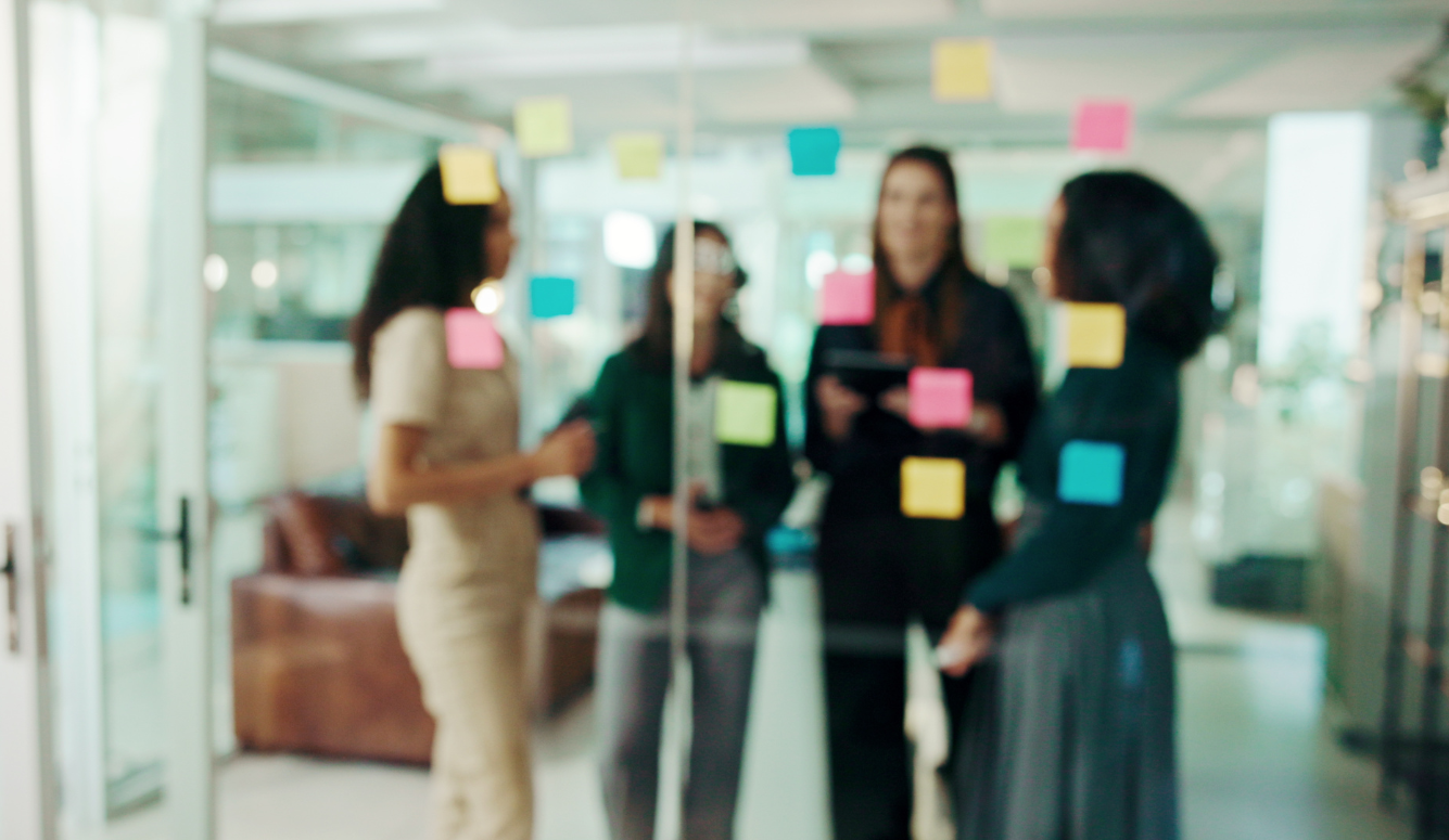 Four women collaborate around a glass wall covered in colourful sticky notes in a modern office setting.