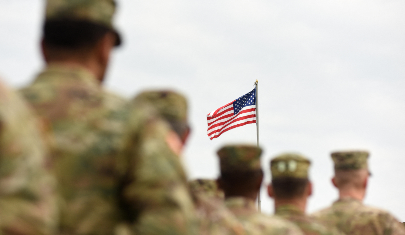 Soldiers in camouflage stand at attention as an American flag waves on a pole.