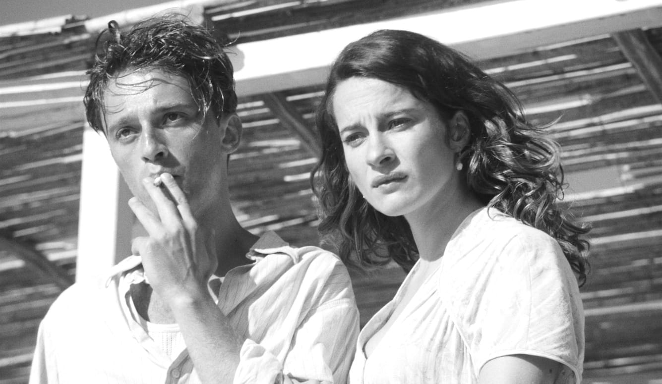 Black-and-white film still: a young man smokes a cigarette while a dark-haired woman beside him gazes ahead with a troubled expression. Slatted structure behind them.