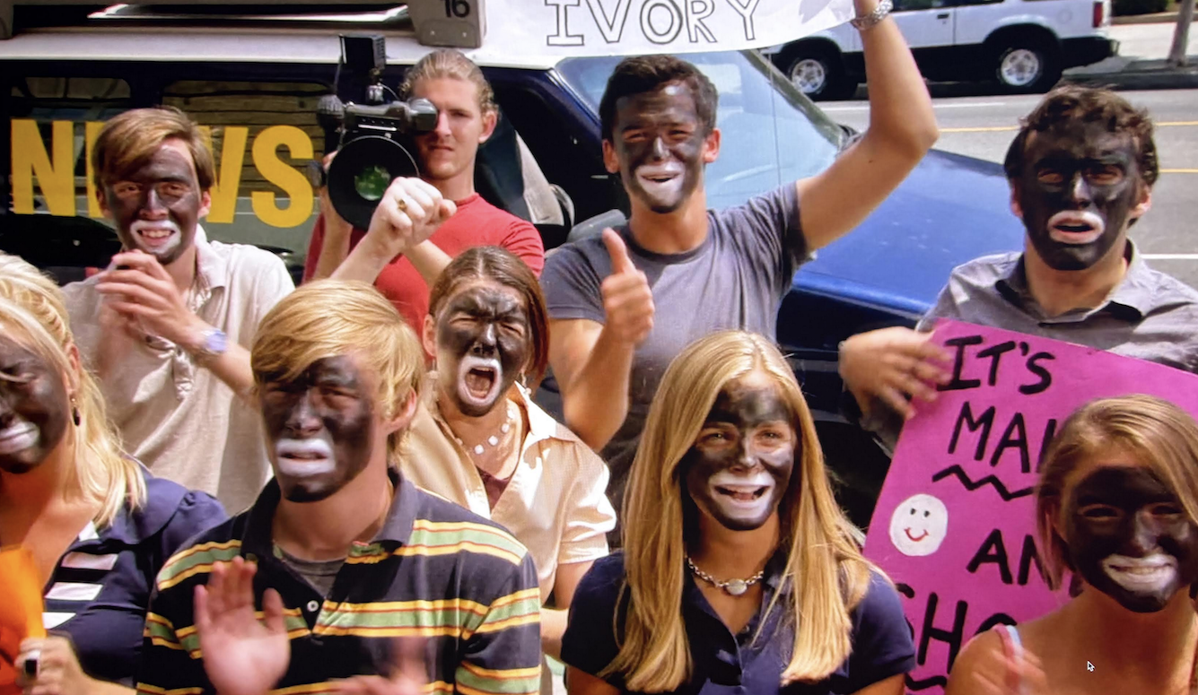A crowd of young people with blackface makeup at an outdoor protest or demonstration, holding signs, with a news camera visible in the background.