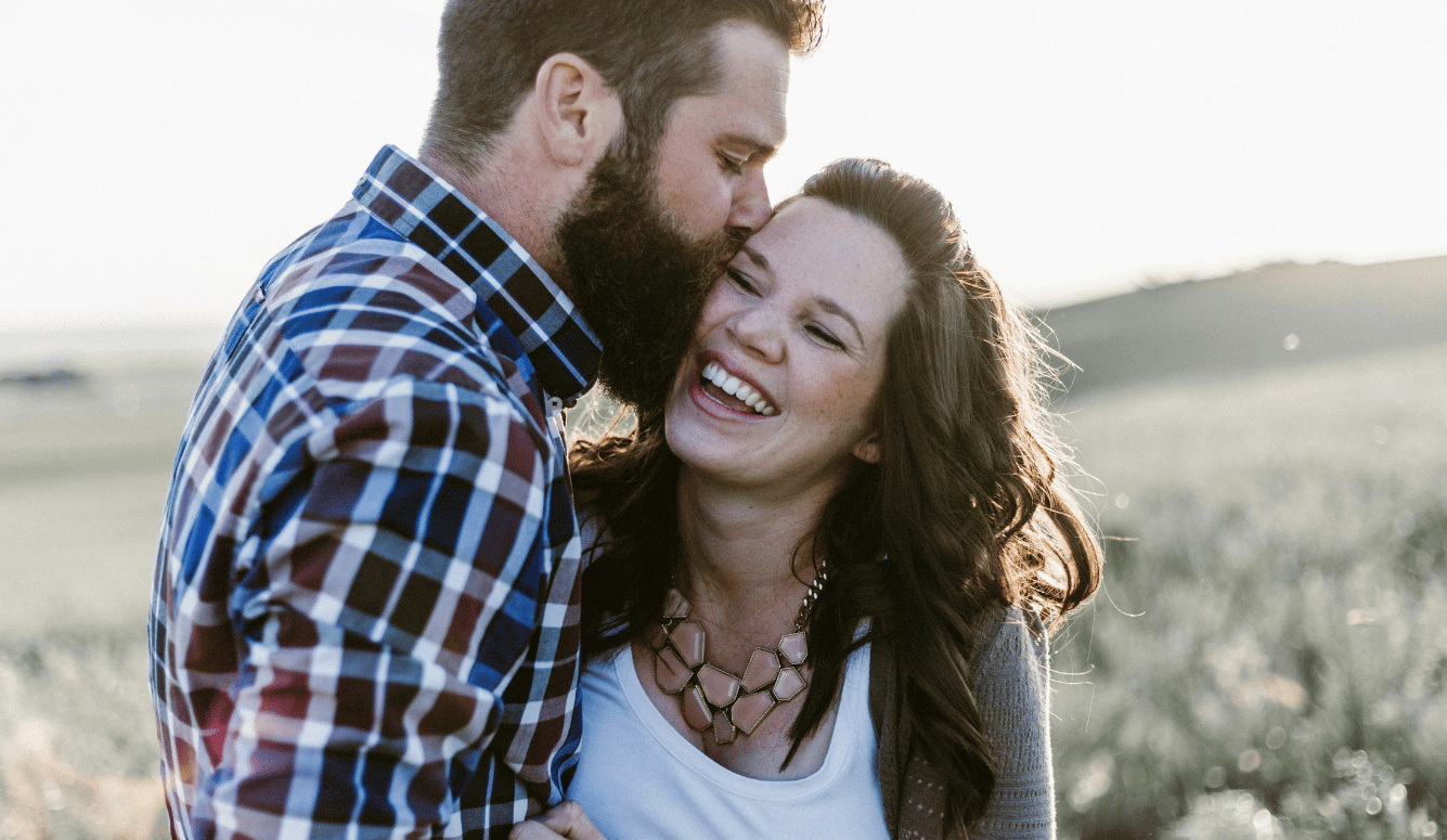 A man in a checked shirt kisses a woman on the cheek. She is hugging him and laughing.