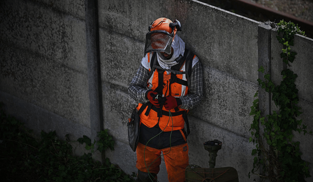 A worker wearing high-visibility orange safety gear, a helmet, and a face shield stands leaning against a concrete wall while looking down."