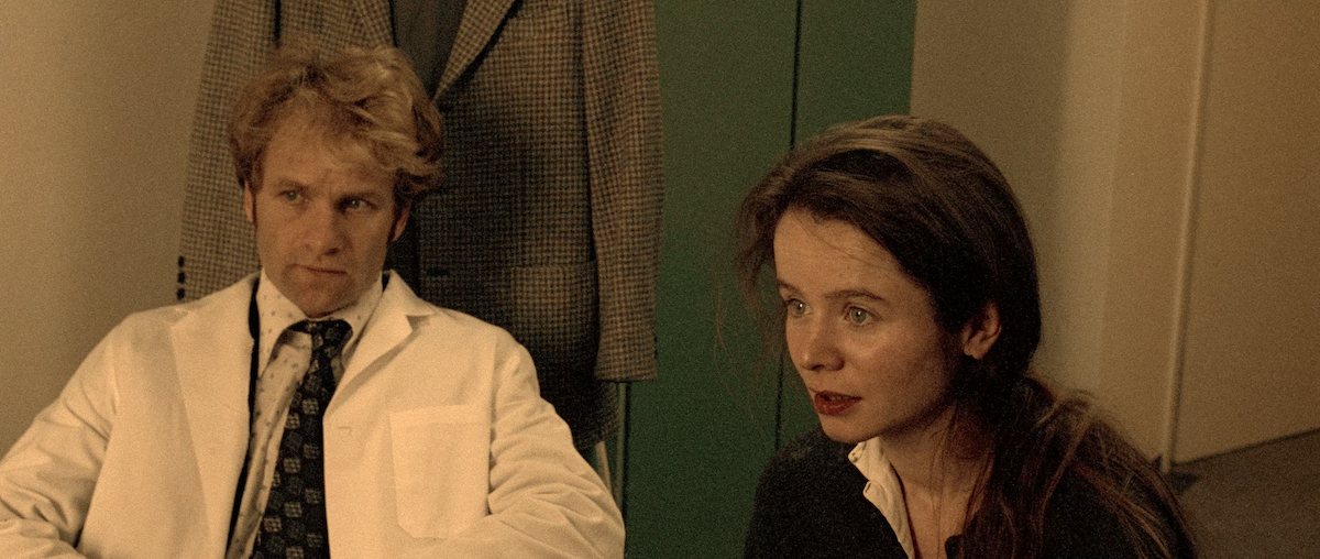 Man in a white doctor's coat sits beside a young woman with dark hair in a ponytail in a hospital consultation room.