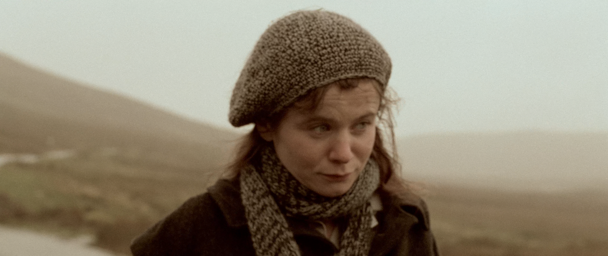 Young woman wearing a knitted beret, scarf and dark coat stands outdoors against a misty rural landscape.