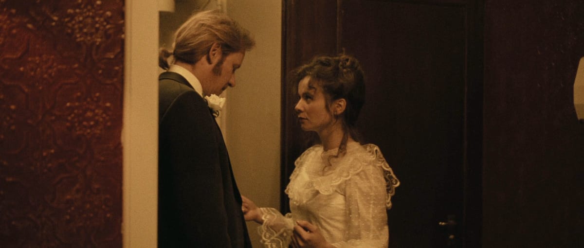 Man in a dark suit and woman in a white lace dress face each other in a dimly lit doorway.