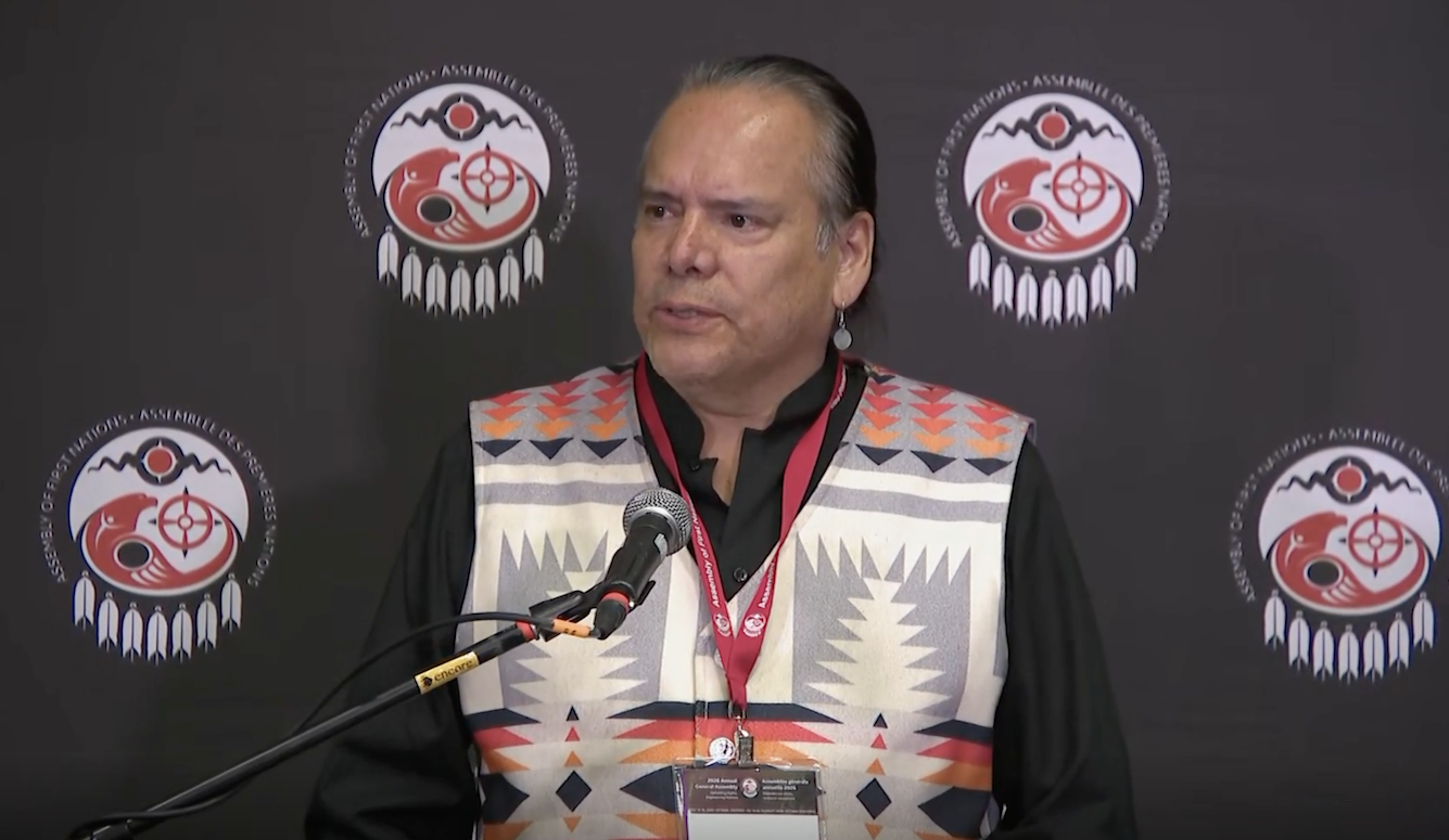 **Alt text (≤125 characters):** Man with long tied-back hair wearing a patterned vest speaks at a microphone before an Assembly of First Nations backdrop.