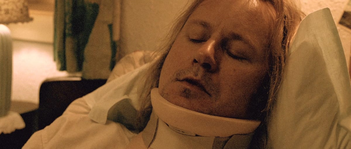Man with shoulder-length blond hair lies in bed wearing a neck brace with his eyes closed in a dimly lit room.