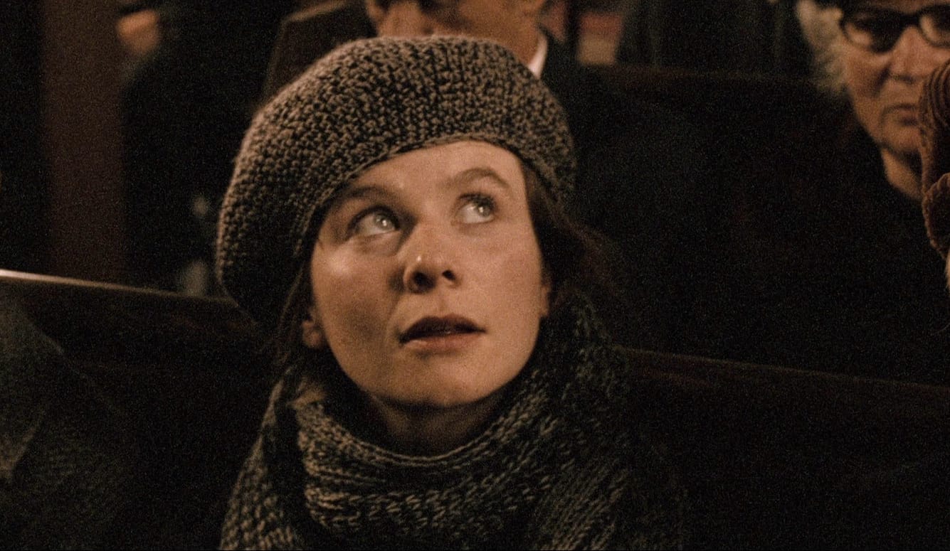 Young woman wearing a knitted beret and scarf looks upward while seated indoors among a crowd.