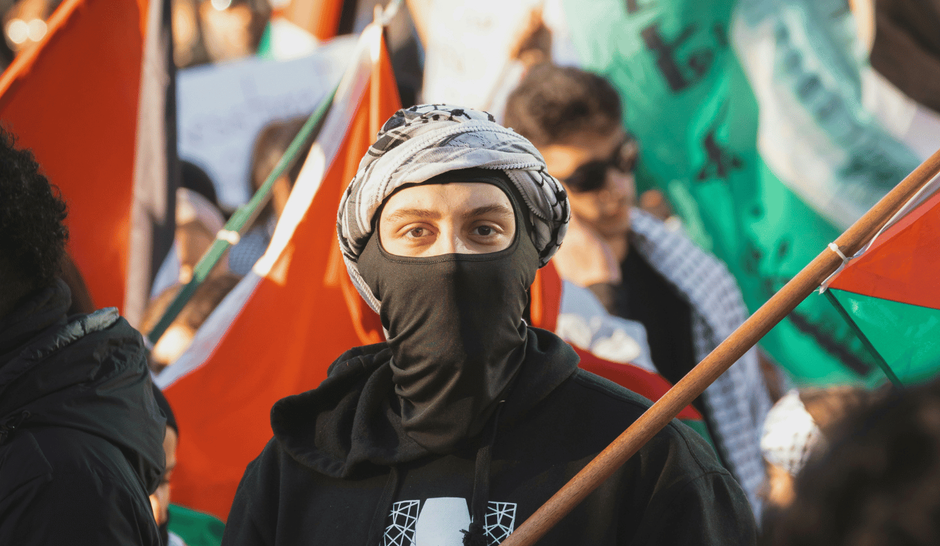 Person in a black hoodie with face covering and headscarf holds a Palestinian flag amid a crowd at a protest.
