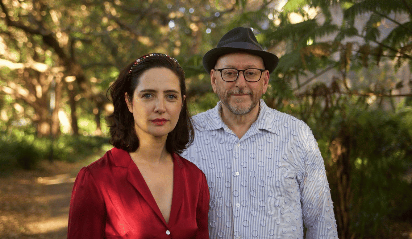 Anna, with shoulder-length dark hair and a decorative headband, wearing a red blouse, stands beside Joseph, who wears glasses, a black hat, a short grey beard in a leafy outdoor setting.