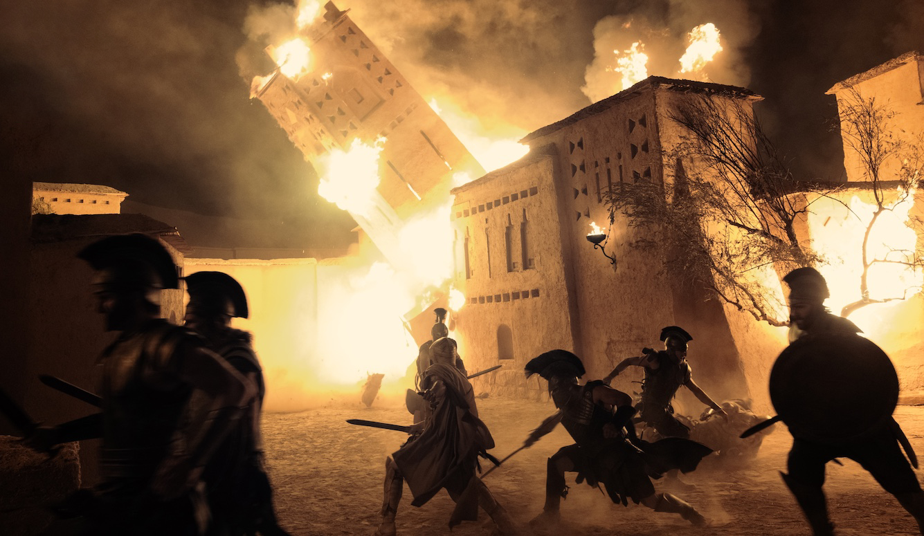 Armoured warriors run through a burning ancient city as flames engulf collapsing buildings in a night battle scene.