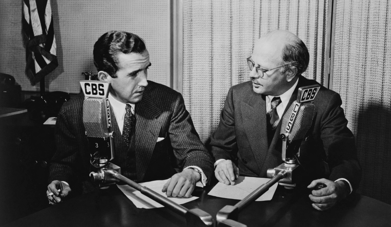 Old photo. Two suited men sit at a desk in a radio studio, facing each other beside CBS microphones and papers; one wears glasses.