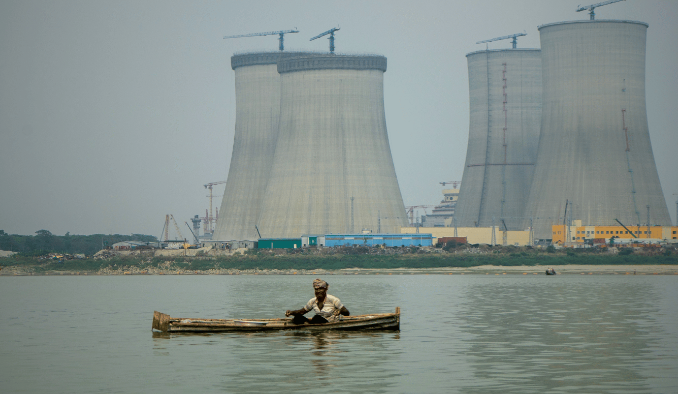 Fisherman in a wooden boat on the Padma River with Rooppur nuclear plant cooling towers behind him.